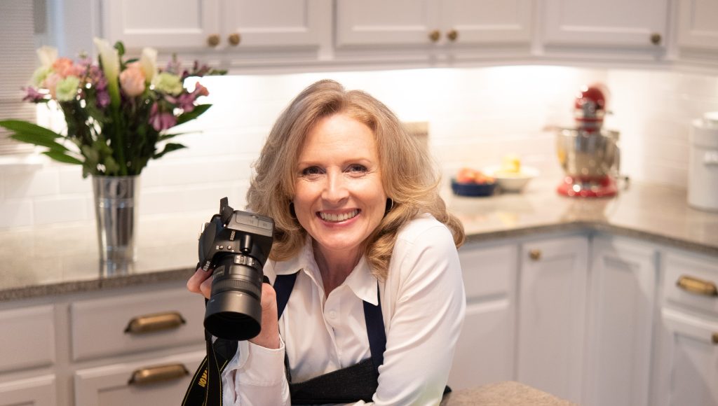 A blonde woman leans over a kitchen counter, holding a Nikon camera. She is wearing a crisp white shirt and a dark apron.